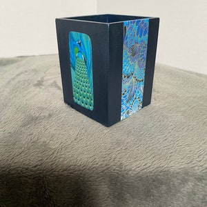 Puede incluir: Un contenedor cuadrado negro con un diseño de pavo real en un lado y un patrón floral azul y dorado en el otro.