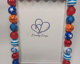 May include: A white picture frame decorated with red, blue, and white beads. The beads feature basketballs, stars, and the Philadelphia 76ers logo. The frame has a white mat and a clear glass front.