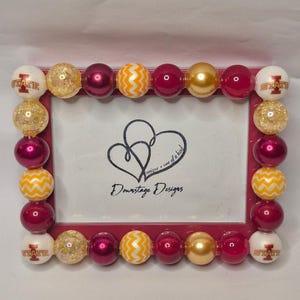 May include: A rectangular red picture frame decorated with large, colorful beads. The beads are white with "STATE" printed on them, gold glitter, maroon, gold, and yellow with a chevron pattern. The frame has the text "Downstage Designs" and a heart design.
