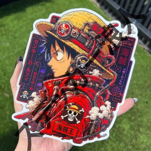 Puede incluir: Una pegatina con un personaje con sombrero de paja y chaqueta roja. Tiene texto japonés y un logotipo de calavera. El personaje es "Monkey D. Luffy". La pegatina tiene un diseño steampunk.