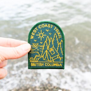 May include: A green and yellow embroidered patch with the text "WEST COAST TRAIL" and "BRITISH COLUMBIA". The patch features a sun, trees, a tent, and a bear. The patch is held up against a blurred background of water and pebbles.