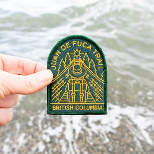 May include: A green and gold embroidered patch with the text "JUAN DE FUCA TRAIL" and "BRITISH COLUMBIA". The patch features a backpack, trees, and a sun design. The patch is held up against a blurred water background.