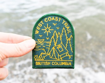 May include: A green and yellow embroidered patch with the text "WEST COAST TRAIL" and "BRITISH COLUMBIA". The patch features a sun, trees, a tent, and a bear. The patch is held up against a blurred background of water and pebbles.