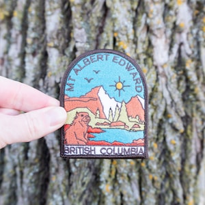 May include: A rectangular patch with a brown border featuring a mountain landscape with the text "MT ALBERT EDWARD" and "BRITISH COLUMBIA". The patch has a blue sky, a sun, and a bear.