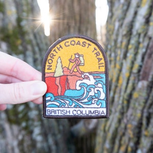 May include: Embroidered patch with a design featuring a hiker on a trail, waves, and the text "North Coast Trail" and "British Columbia." The patch has a brown border, yellow, orange, red, and blue colours.