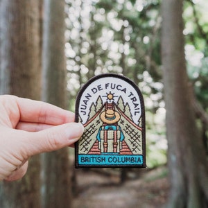 May include: A rectangular patch with the text "JUAN DE FUCA TRAIL" and "BRITISH COLUMBIA". The patch features a hiker with a backpack, a hat, and a sun graphic. The patch is held in front of a blurred forest background.