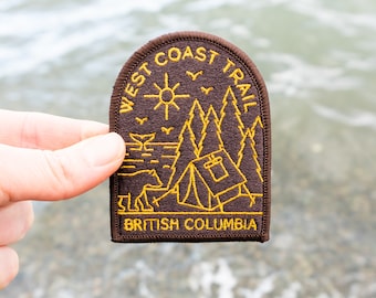 May include: A brown and gold embroidered patch with the text "WEST COAST TRAIL" and "BRITISH COLUMBIA". The patch features a sun, trees, a tent, and a whale. The patch is held up against a blurred water background.