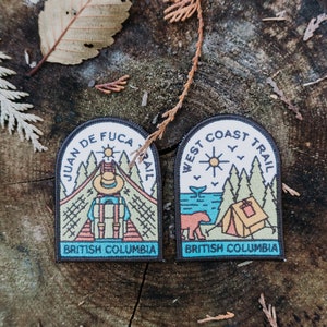 Może przedstawiać: Dwie haftowane naszywki o tematyce outdoorowej. Jedna głosi "Juan de Fuca Trail", a druga "West Coast Trail", obie z napisem "British Columbia" na dole. Naszywki przedstawiają ilustracje gór, drzew i namiotu.
