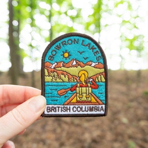 May include: Embroidered patch with the text "BOWRON LAKES BRITISH COLUMBIA". The patch shows a lake, mountains, sun, and a person in a canoe. The patch is held in a hand. The patch is a travel souvenir.