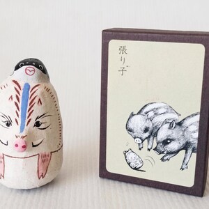 Peut inclure: Une figurine artisanale de cochon blanc cassé avec des détails noirs, bleus et roses. À côté, une boîte marron avec un dessin de marcassins et du texte japonais.