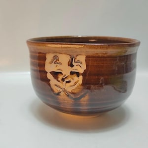 Puede incluir: Un cuenco de cerámica marrón con acabado brillante. El cuenco presenta un diseño tallado de máscaras teatrales en la parte delantera. El cuenco tiene una forma ancha y redondeada y es adecuado para servir o exhibir.