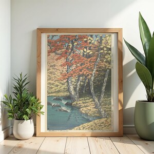 May include: A framed art print showing a tranquil forest scene with a river. The trees have striking red and orange leaves, set within a wooden frame. The artwork is displayed on a light wood floor.