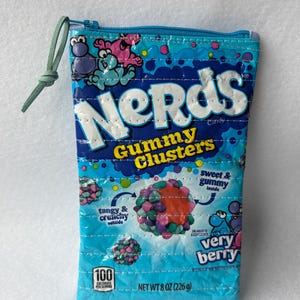 May include: A rectangular, zippered pouch made from a Nerds Gummy Clusters candy package. The pouch is primarily blue with the candy's logo and graphics. It features a green cord zipper pull and is 8 oz (226 g).