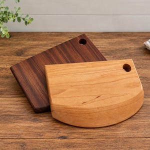 May include: Two wooden cutting boards, one rectangular and dark brown, the other semi-circular and light tan. Both boards feature a circular hole near the top corner for hanging or handling, showcasing natural wood grain patterns and smooth surfaces.