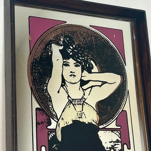 May include: Art Nouveau style mirror with a dark wood frame. The mirror features a stylized illustration of a woman with long hair, set against a pink and gold background. The woman is wearing a cream-colored dress and black dress.