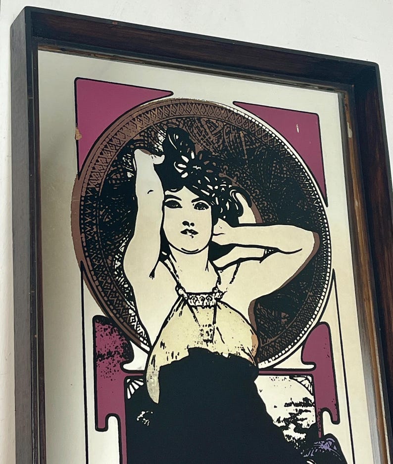 May include: Art Nouveau style mirror with a dark wood frame. The mirror features a stylized illustration of a woman with long hair, set against a pink and gold background. The woman is wearing a cream-colored dress and black dress.