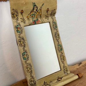 May include: A decorative mirror with a scroll-like frame. The frame is painted with a floral design and a woman playing a musical instrument. The mirror is hanging from a wire.