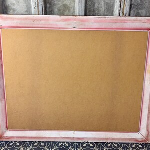 May include: A rectangular picture frame viewed from the back, featuring a light-colored wooden border with distressed pink paint accents. The center is filled with a flat, tan-colored backing board, and two metal hanging brackets are attached.