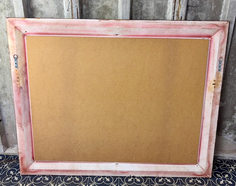 May include: A rectangular picture frame viewed from the back, featuring a light-colored wooden border with distressed pink paint accents. The center is filled with a flat, tan-colored backing board, and two metal hanging brackets are attached.