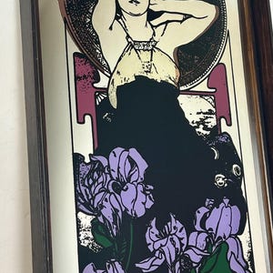 May include: Art Nouveau style print featuring a woman with her arms raised, surrounded by a circular design and purple irises. The woman is wearing a cream-colored top and a black dress. The print is framed in a dark wood frame.