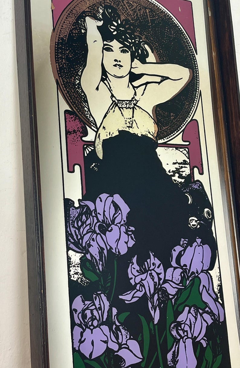 May include: Art Nouveau style print featuring a woman with her arms raised, surrounded by a circular design and purple irises. The woman is wearing a cream-colored top and a black dress. The print is framed in a dark wood frame.