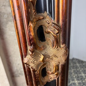 May include: A vertical mirror frame featuring a deep black central channel flanked by raised, burnished gold-toned borders. The frame is adorned with ornate, relief-style gold-colored scrollwork and foliage motifs that extend along the vertical length.