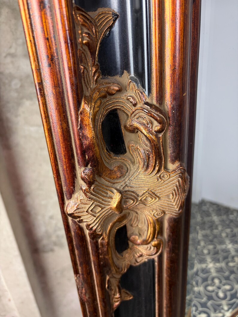 May include: A vertical mirror frame featuring a deep black central channel flanked by raised, burnished gold-toned borders. The frame is adorned with ornate, relief-style gold-colored scrollwork and foliage motifs that extend along the vertical length.