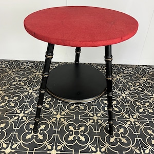 Könnte beinhalten: Ein schwarzer und roter zweistufiger Beistelltisch mit runder Tischplatte und drei Beinen. Die obere Stufe ist mit rotem Stoff bezogen, die untere Stufe ist schwarz.