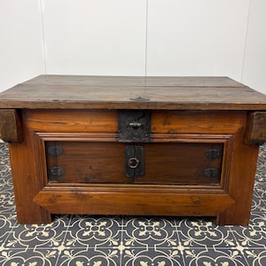 May include: A rectangular wooden chest with a dark brown finish, featuring a flat top surface, a central front panel with two small doors, and dark metal hardware including hinges, a latch, and a circular pull ring on the front face.
