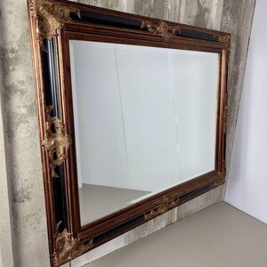 May include: A rectangular wall mirror featuring a wide, ornate frame with a combination of dark black and metallic bronze finishes. The frame is decorated with intricate, raised scrollwork and floral-inspired carvings at each corner and edge.