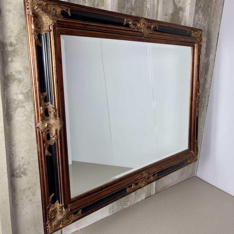 May include: A rectangular wall mirror featuring a wide, ornate frame with a combination of dark black and metallic bronze finishes. The frame is decorated with intricate, raised scrollwork and floral-inspired carvings at each corner and edge.