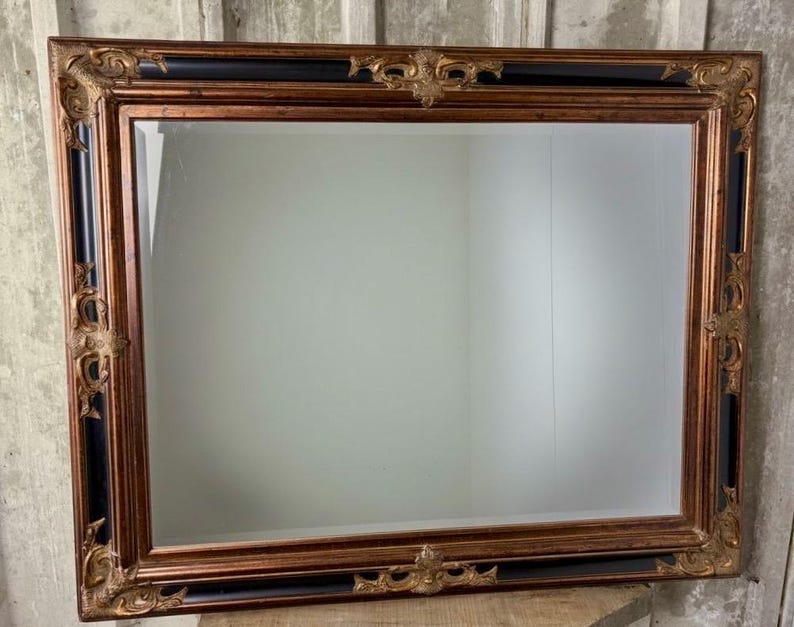 May include: A rectangular wall mirror featuring a wide, ornate frame with a combination of metallic bronze and deep black finishes. The frame is decorated with intricate, raised scrollwork and floral motifs at the corners and center edges.
