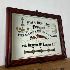 Puede incluir: Espejo rectangular vintage con marco de madera oscura. La superficie del espejo presenta texto e ilustraciones en dorado y rojo, anunciando "JOHN ROGERS, Pewterers, Beer Engine & Pewter Pot Makers, Gas Fitters &c" con una dirección en Londres.