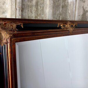 May include: A large rectangular mirror featuring a wide wooden frame with a rich brown finish. The frame is accented by ornate, raised gold-toned decorative corner pieces and a central top embellishment, set against a smooth black inner border.