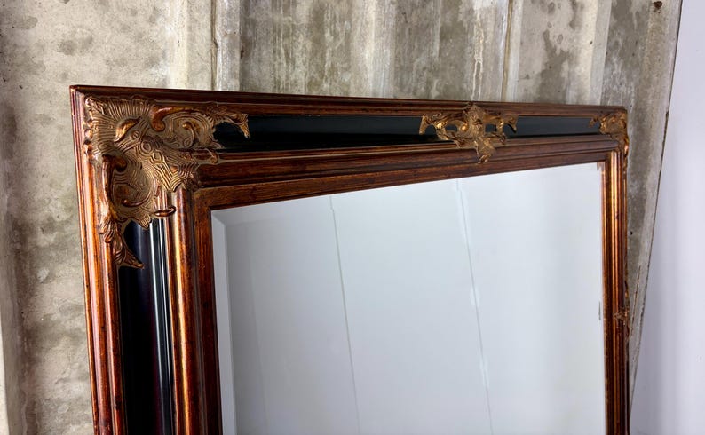 May include: A large rectangular mirror featuring a wide wooden frame with a rich brown finish. The frame is accented by ornate, raised gold-toned decorative corner pieces and a central top embellishment, set against a smooth black inner border.