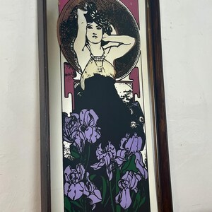May include: Art Nouveau style print in a dark wood frame. The image features a woman with her arms raised, surrounded by a circular design. Below the woman is a cluster of purple irises against a black background. The frame is rectangular.