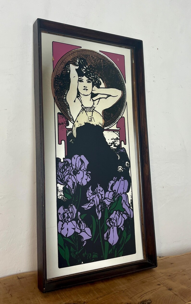 May include: Art Nouveau style print in a dark wood frame. The image features a woman with her arms raised, surrounded by a circular design. Below the woman is a cluster of purple irises against a black background. The frame is rectangular.