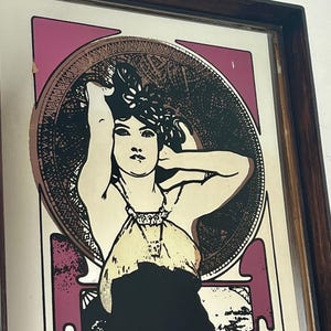 May include: Framed vintage art nouveau print featuring a woman with long hair, wearing a dress and jewelry. The artwork has a color palette of black, cream, purple, and gold. The print is in a dark brown wooden frame.