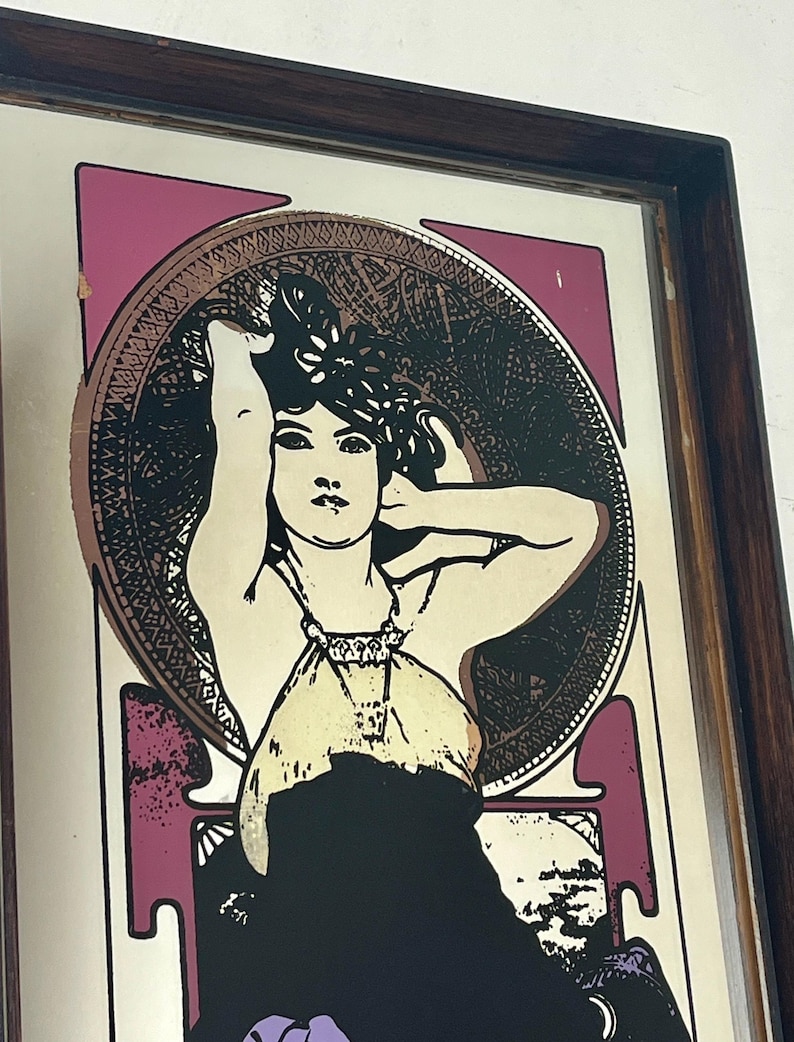 May include: Framed vintage art nouveau print featuring a woman with long hair, wearing a dress and jewelry. The artwork has a color palette of black, cream, purple, and gold. The print is in a dark brown wooden frame.