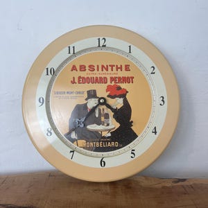 Puede incluir: Un reloj vintage con una esfera amarilla y números negros. El reloj presenta un anuncio vintage de Absinthe Extra-Superieure J. Edouard Pernot. El anuncio muestra a un hombre y una mujer sentados a una mesa con una botella de absenta.