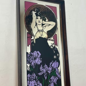 May include: Rectangular framed art print featuring a woman with her arms raised, surrounded by a circular design and purple irises. The artwork has a dark brown frame and a color palette of black, purple, and pink.