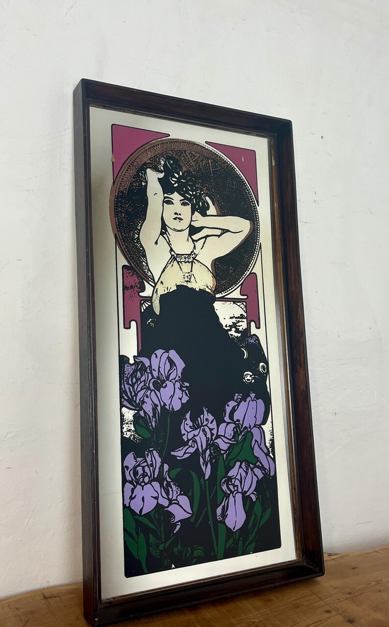 May include: Rectangular framed art print featuring a woman with her arms raised, surrounded by a circular design and purple irises. The artwork has a dark brown frame and a color palette of black, purple, and pink.