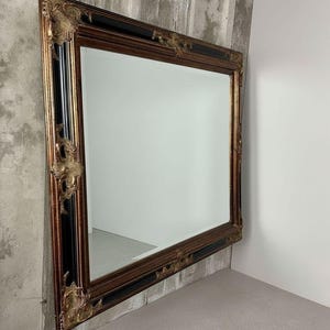 May include: A large rectangular wall mirror featuring a wide frame with a combination of black and metallic gold finishes. The frame is accented with ornate, raised gold-toned decorative corner and side embellishments in a relief pattern.