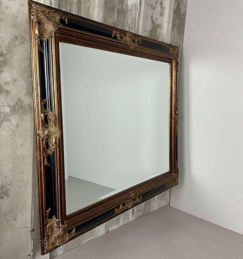 May include: A large rectangular wall mirror featuring a wide frame with a combination of black and metallic gold finishes. The frame is accented with ornate, raised gold-toned decorative corner and side embellishments in a relief pattern.