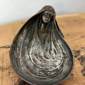 A drop-shaped metal bowl with a woman's head as a detail. The bowl is in art nouveau style, and the woman has long hair and a dreamy face—the bowl is a mixture of different tones of silver grey and sins of vintage patina.