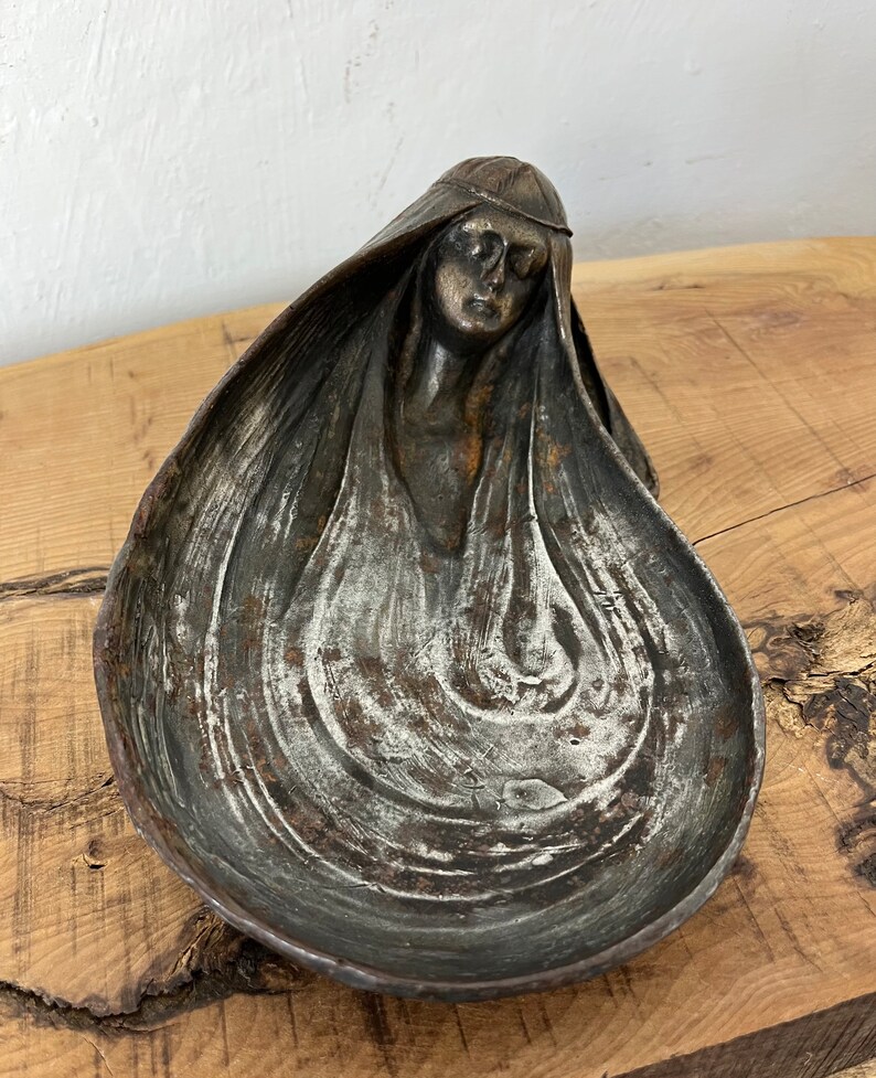 A drop-shaped metal bowl with a woman's head as a detail. The bowl is in art nouveau style, and the woman has long hair and a dreamy face—the bowl is a mixture of different tones of silver grey and sins of vintage patina.