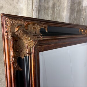 May include: A rectangular mirror featuring a dark brown wooden frame with a glossy black inner border. The corners are adorned with ornate, raised metallic gold-toned decorative accents that feature intricate, swirling relief patterns.