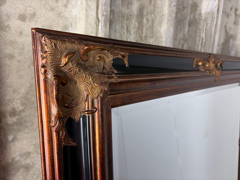 May include: A rectangular mirror featuring a dark brown wooden frame with a glossy black inner border. The corners are adorned with ornate, raised metallic gold-toned decorative accents that feature intricate, swirling relief patterns.