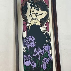 May include: Art Nouveau style framed print featuring a woman with her arms raised, surrounded by a circular design and purple irises. The artwork has a black background and a dark brown wooden frame.