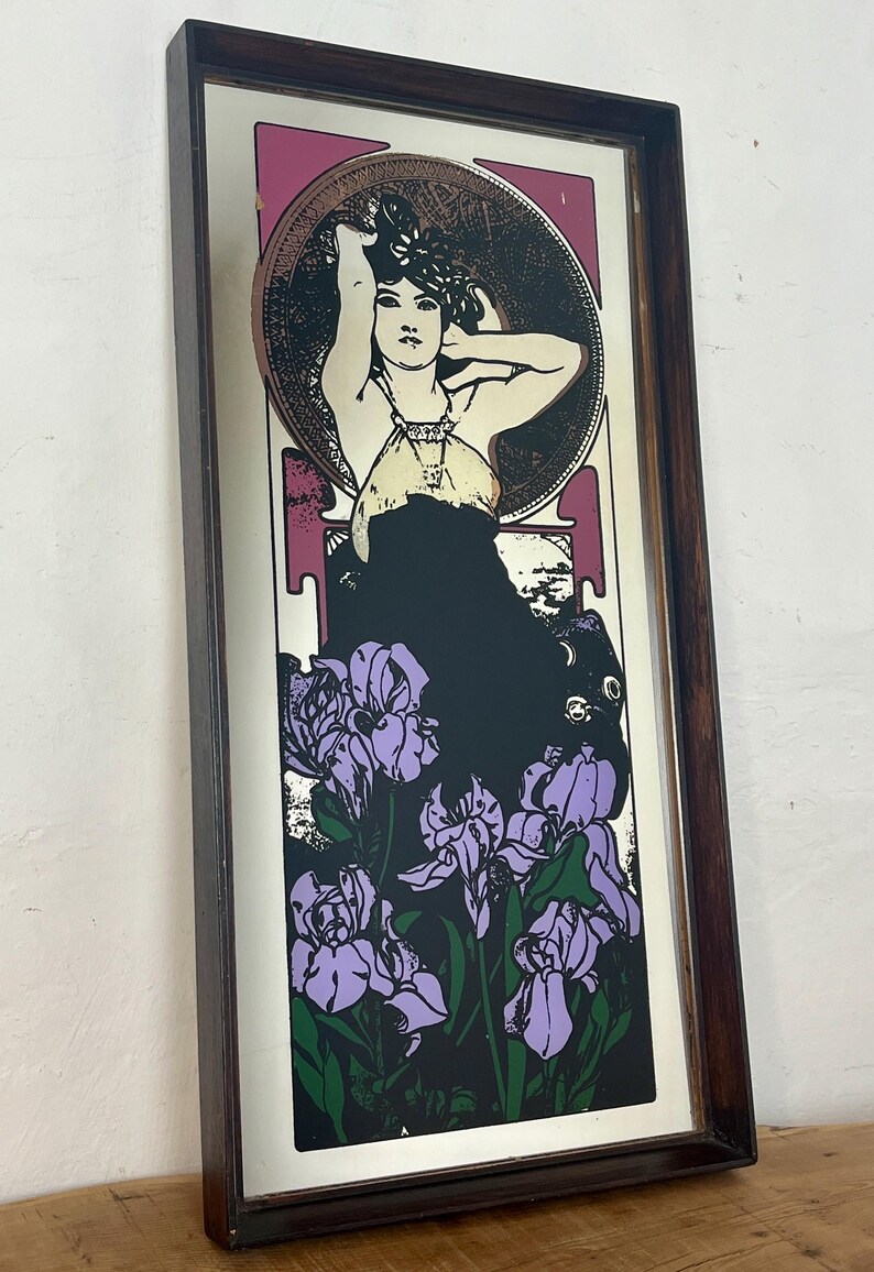 May include: Art Nouveau style framed print featuring a woman with her arms raised, surrounded by a circular design and purple irises. The artwork has a black background and a dark brown wooden frame.